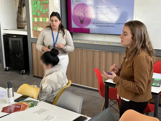 Cote, con credencial, guía un taller frente a una pantalla sobre la fase idear junto a otra profesora; la alumna sentada tiene la cara difuminada